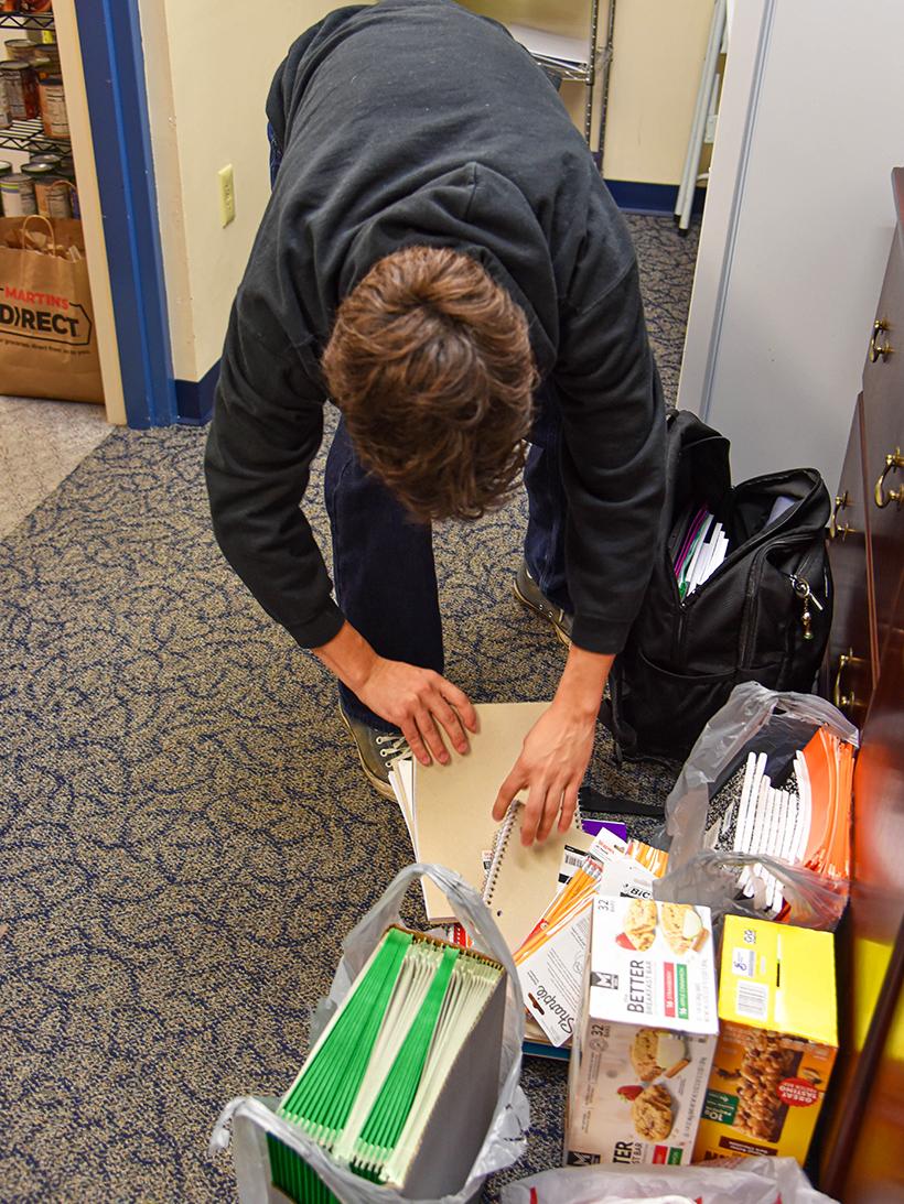 Person organizing donated school supplies and food items into bags inside a room with shelves of canned goods in the background.