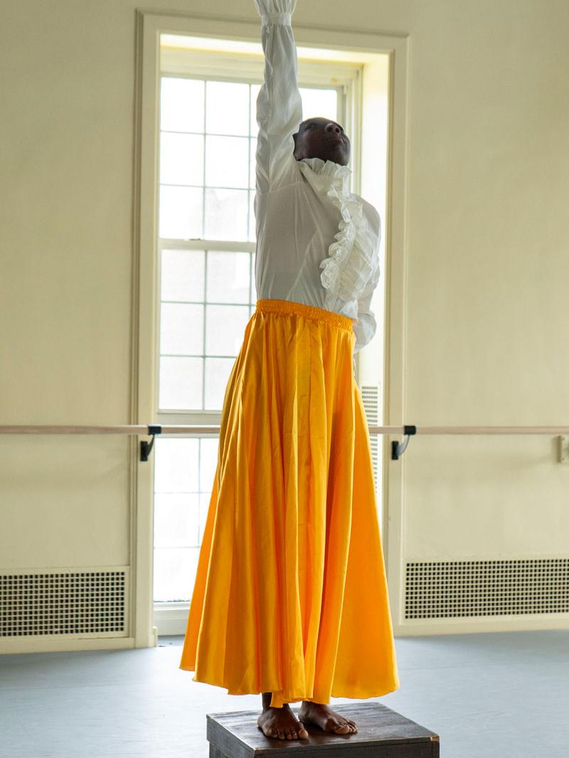 A dancer wearing a long skirt stands on a box and reaches one arm upward.