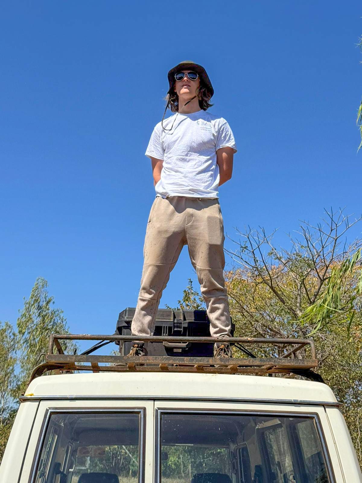 A man in a grey shirt stands on top of a vehicle overlooking a field.