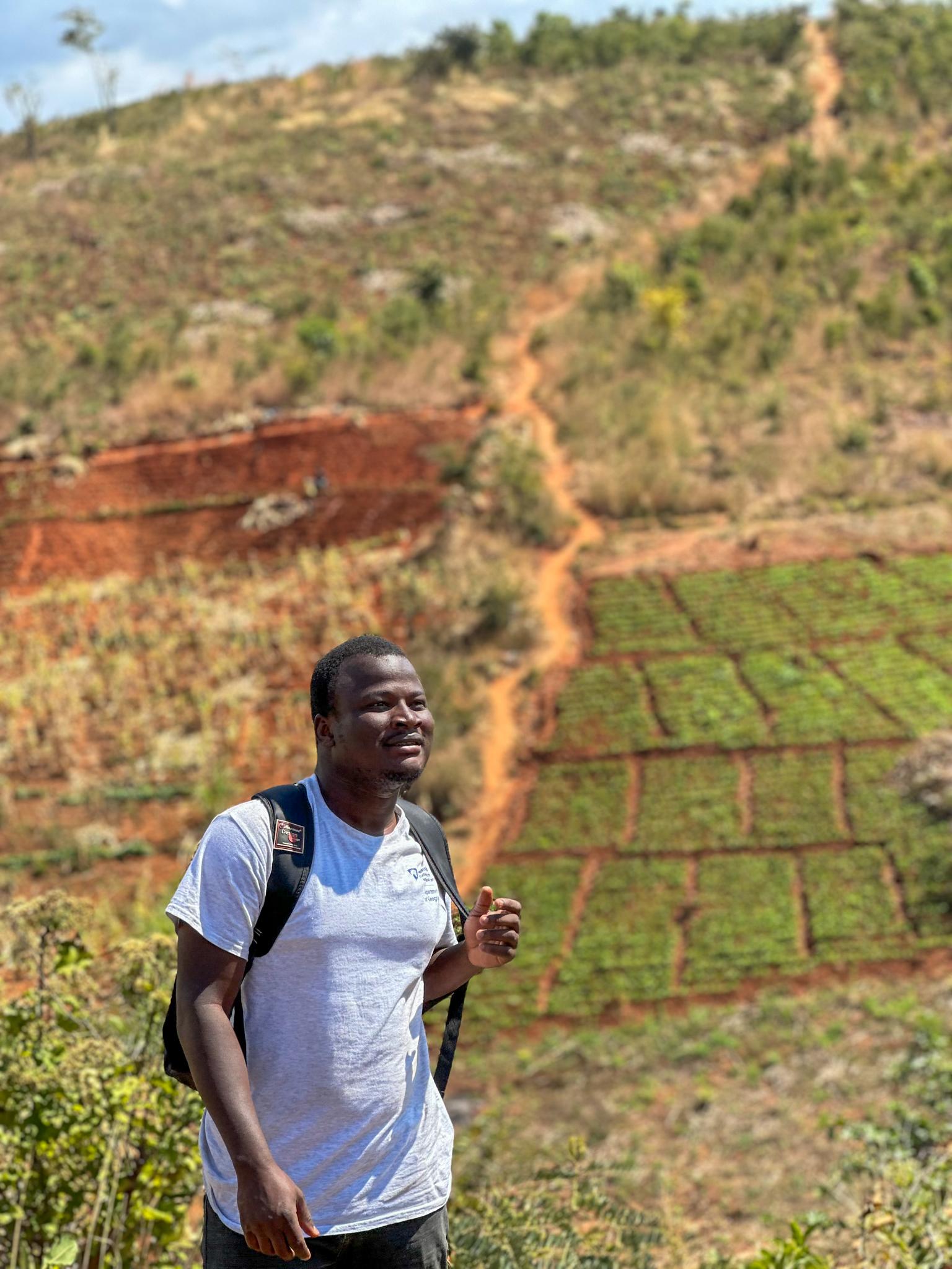 A smiling man in a grey shirt and backpack stands before a terraced hillside with brown and green patches under a blue sky.