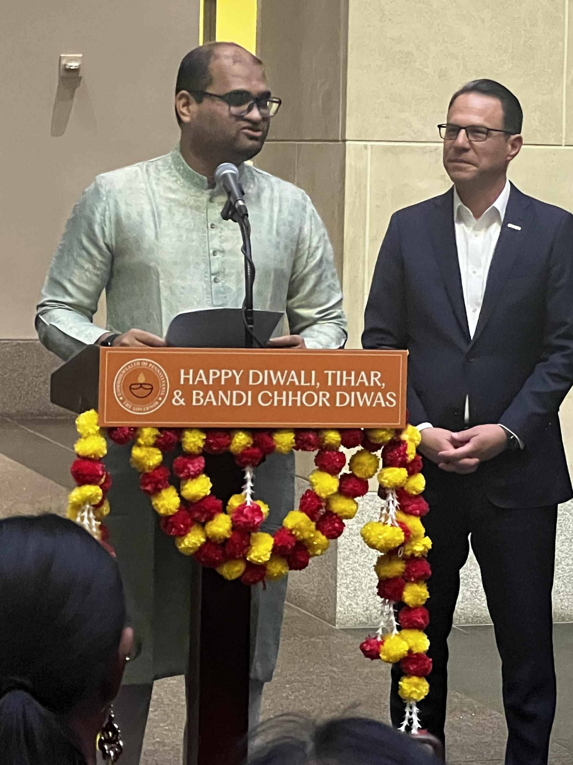 Two men standing in front of a podium.  One is speaking and one is waiting.