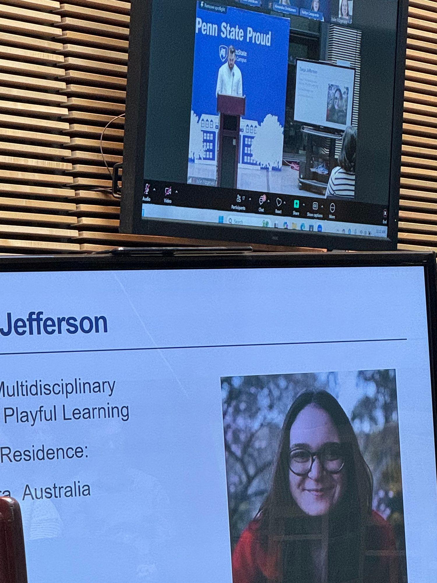 A TV screen in the foreground shows a slide from a presentation about a student who could not attend in person. A screen in the background shows the livestreamed event.