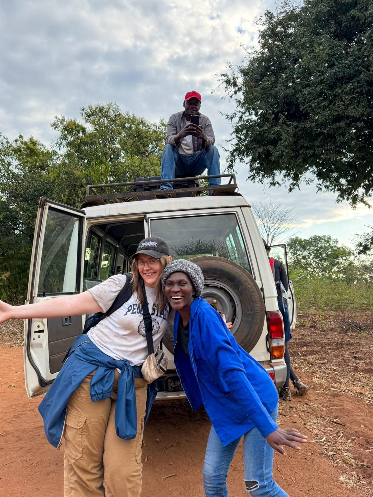 Two women pose together in front of a off-road vehicle. A man sits on top of the car.