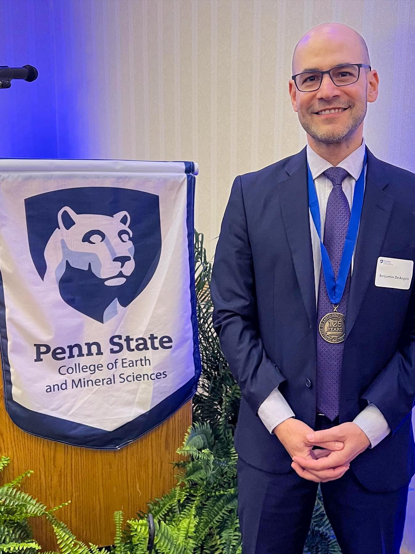 Man in a suit with a medal stands next to a Penn State banner.
