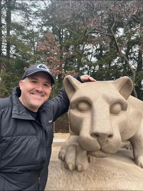 a man in a penn state hat stands next to the nittany lion statue