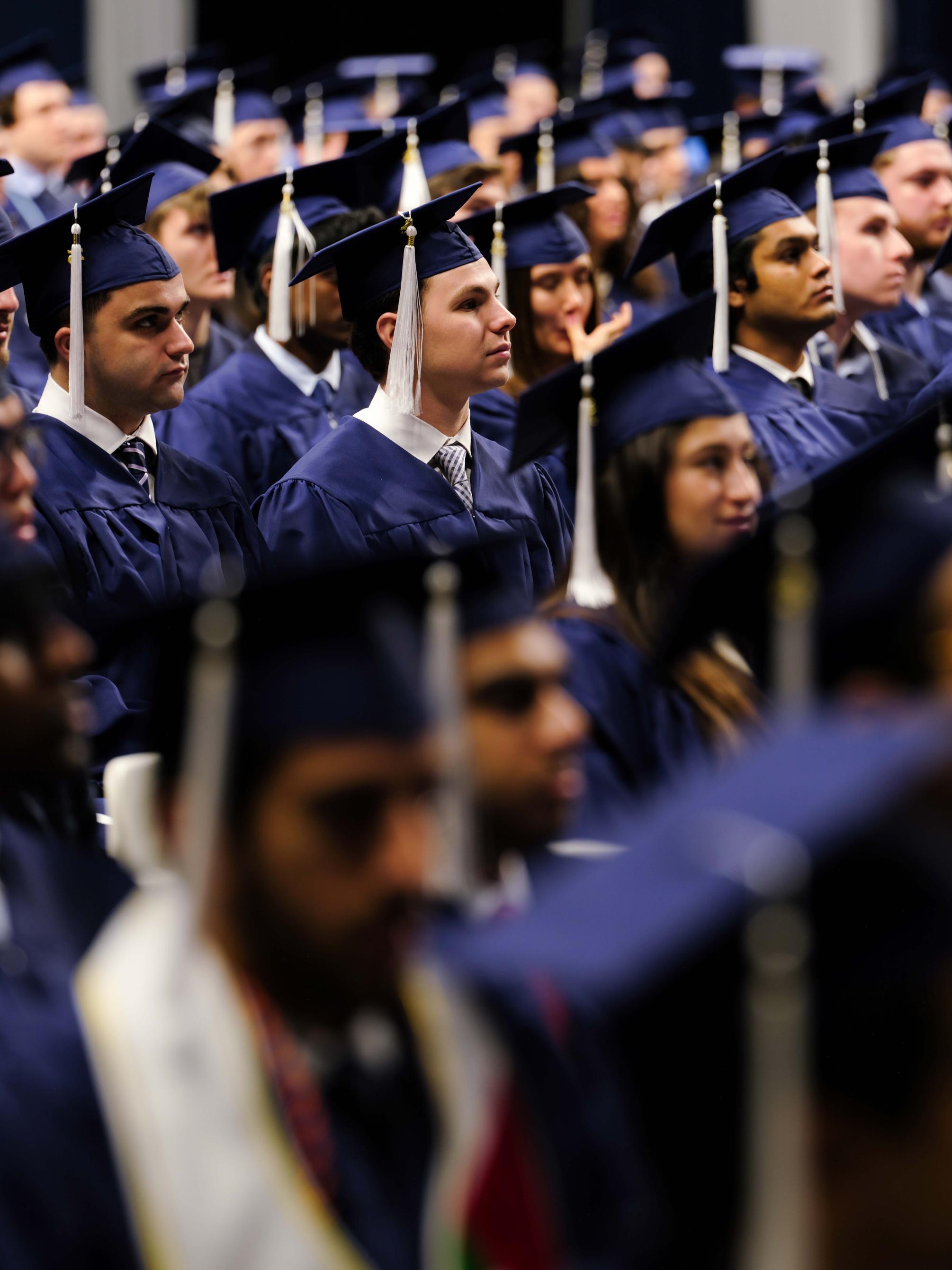 Rows of graduates seated indoors at a commencement ceremony, wearing navy blue caps and gowns with white tassels, facing forward and listening attentively during the program.