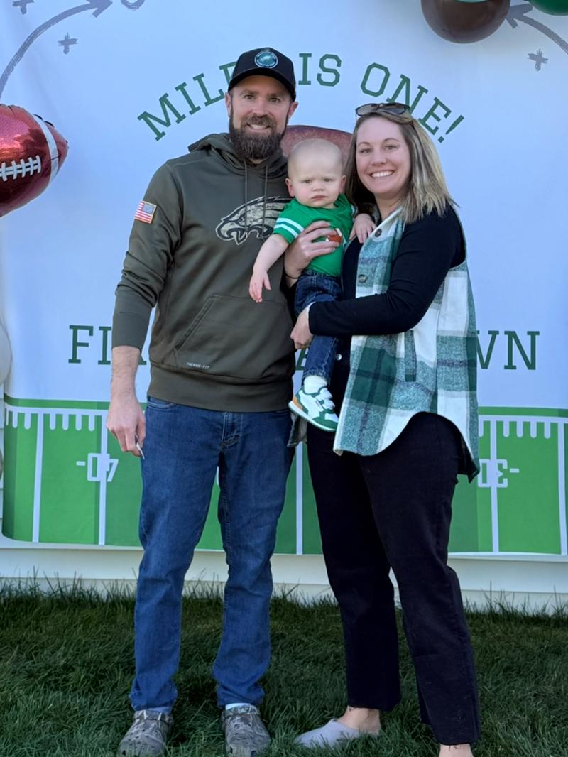 A man and woman hold their baby boy in front of a background that reads “Miles is one!” Balloons surround the background.