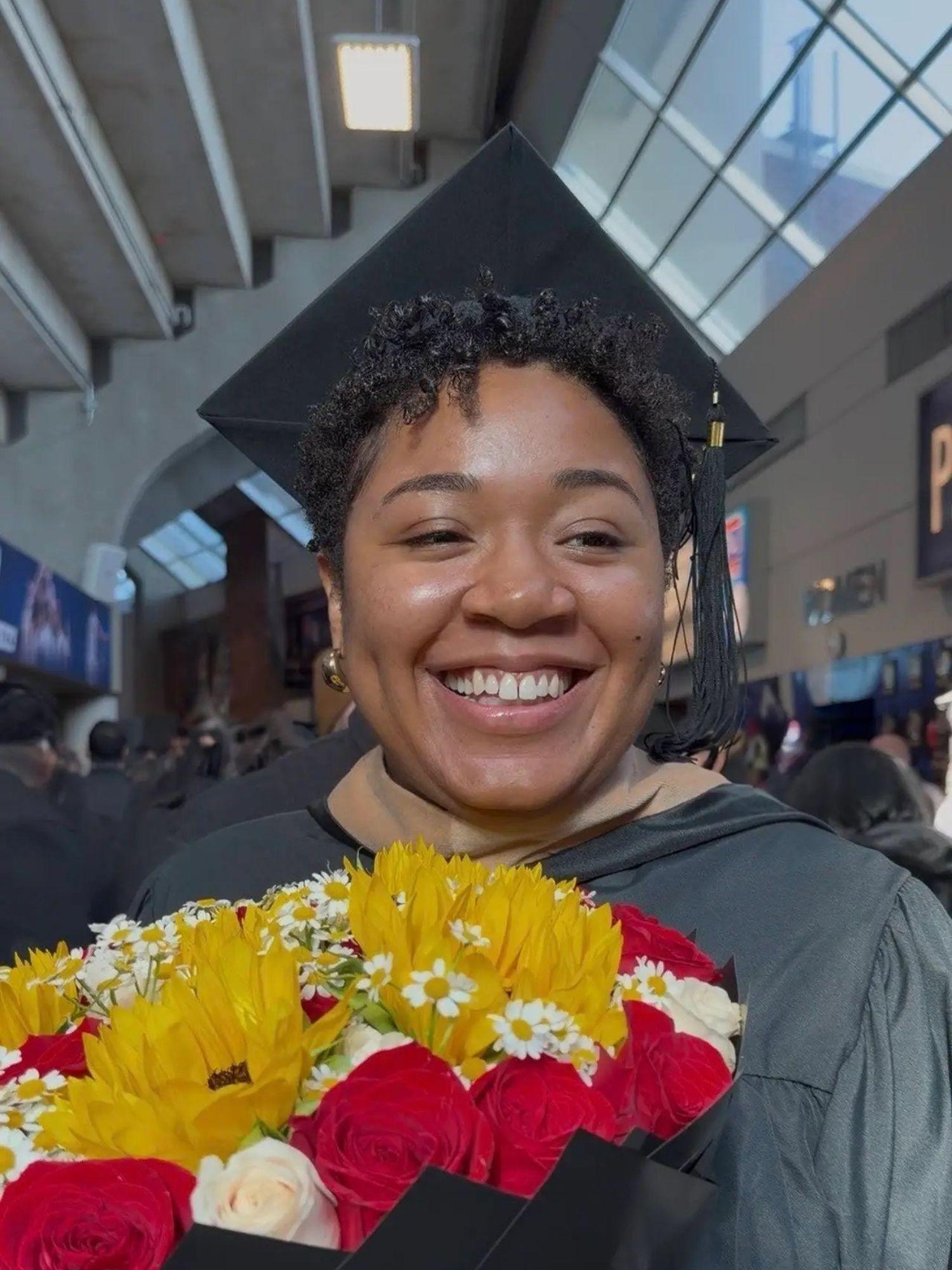 A woman in a commencement cap and gown holds a bouquet of flowers.