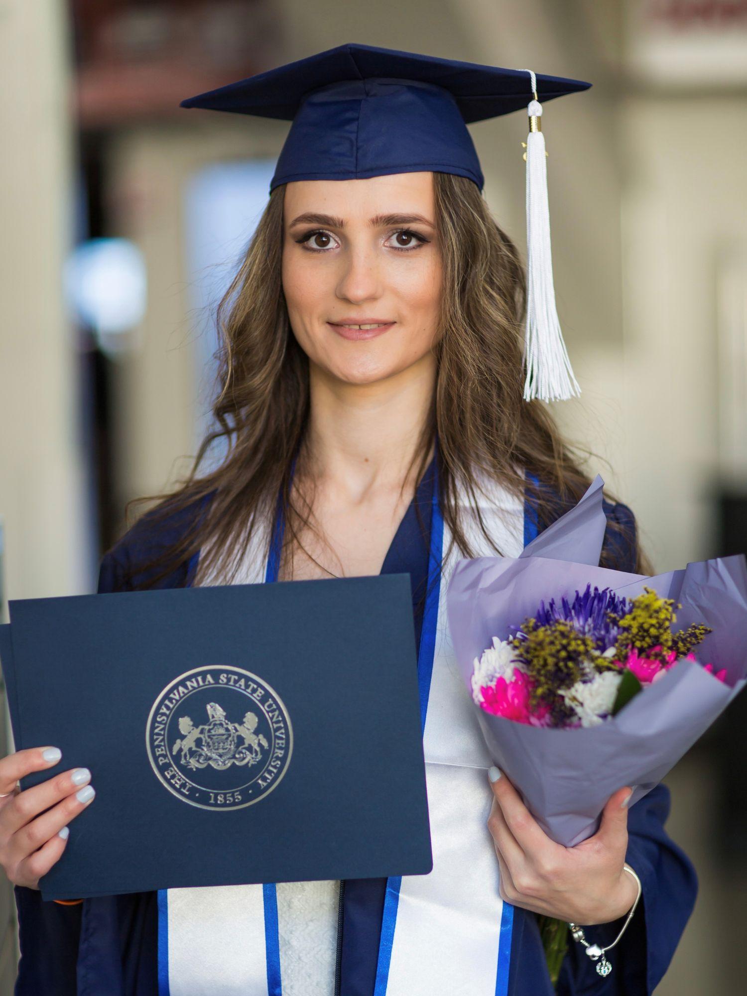 a woman in a graduation cap and gown holds her diploma and a bouquet of flowers.