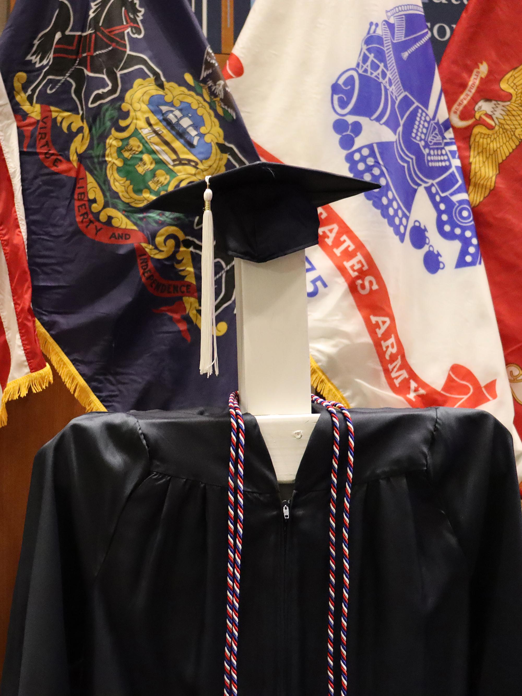 A blue graduation cap and gown with red, white, and blue honor cords in front of a series of U.S. military flags.