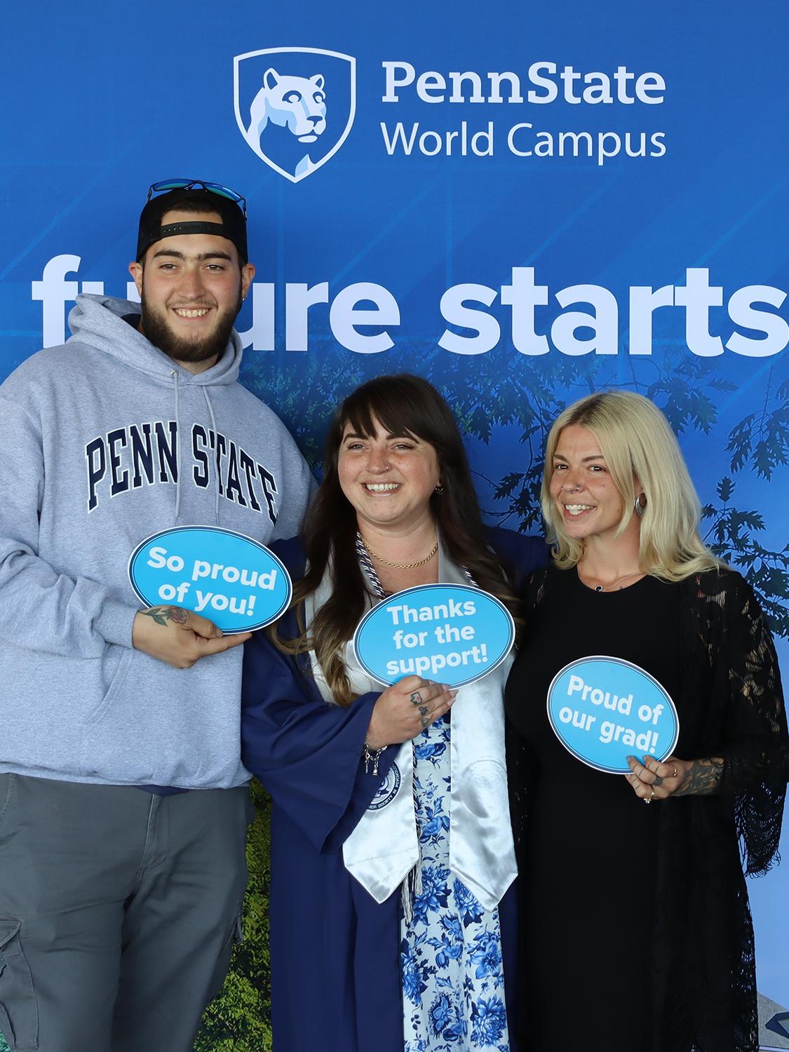 Three people holding signs standing in front of a Penn State photo background.