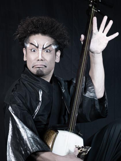 A man wears traditional Japanese costume makeup and holds a stringed instrument.
