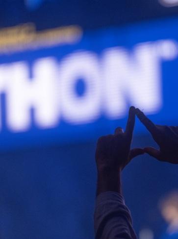 A person in the shadow raises their hands in the shape of a diamond in the foreground of a lit "THON" sign
