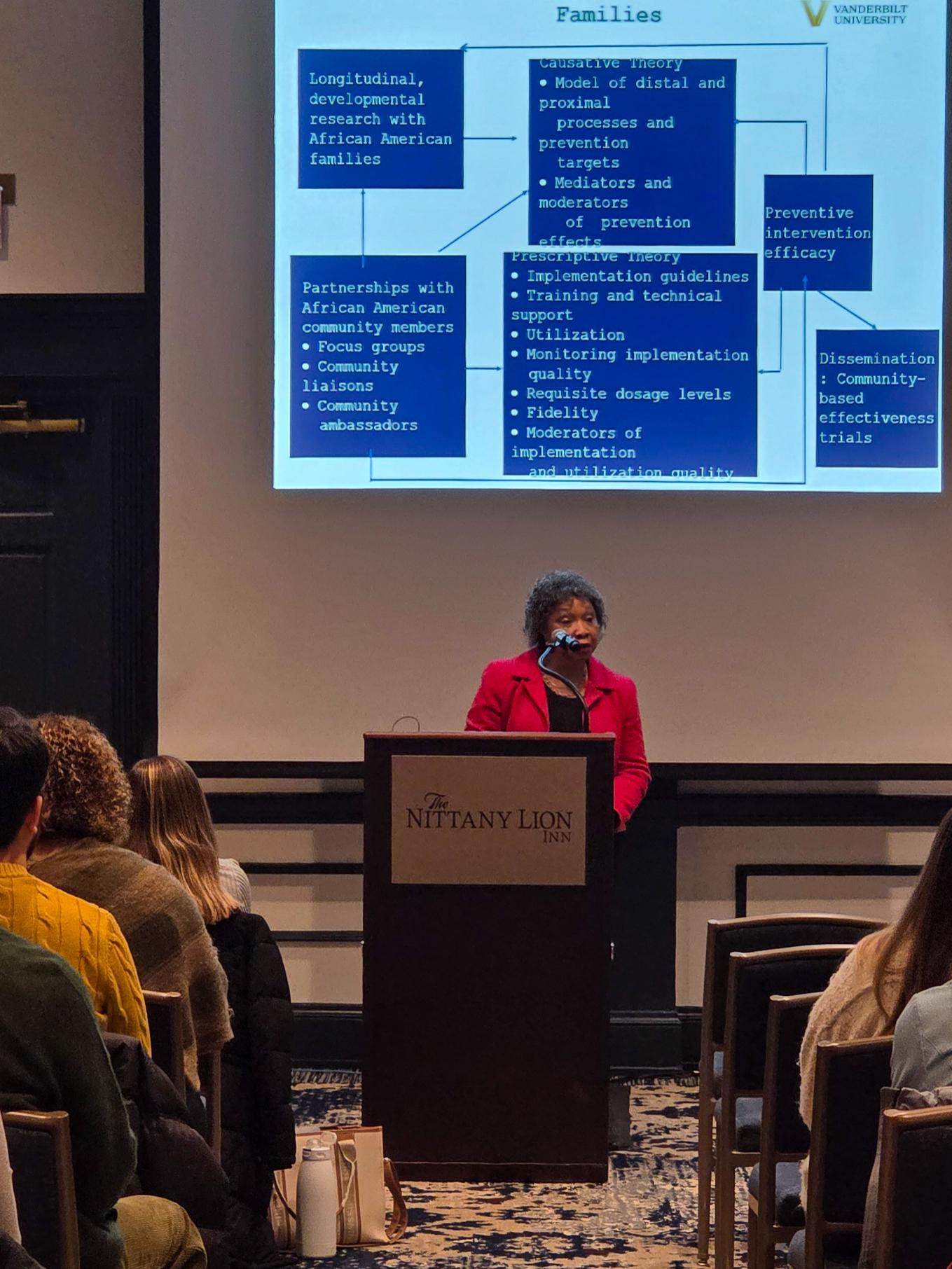 A speaker stands at a podium presenting a slide that shows a diagram of equity indicators and contextual factors, with attendees looking toward the screen.