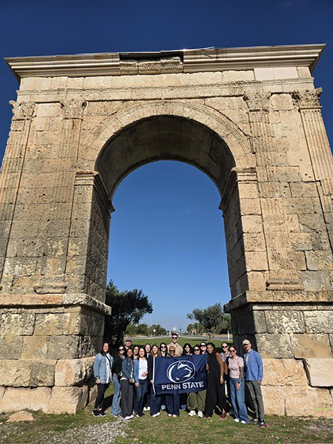 A group of people holding a navy Penn State flag in front of a stone arch.