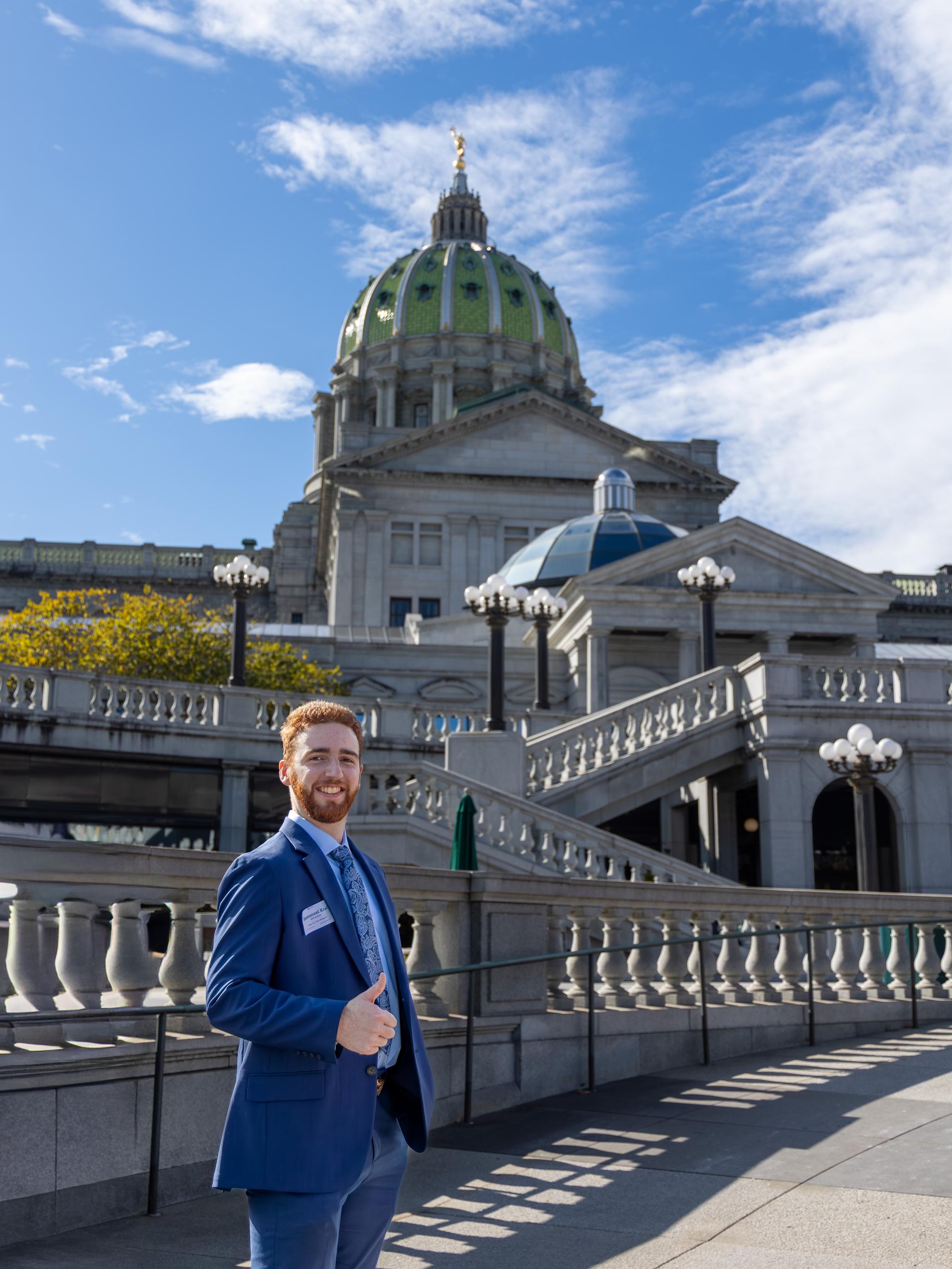 Emmanuel Kraft stands outside the Pennsylvania State Capitol in Harrisburg, wearing a blue suit, smiling and making a thumbs-up gesture while the building’s green dome is visible behind him.