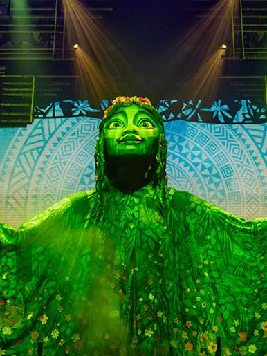 A theatre stage set with a large green goddess being looked at by a woman with brown curly hair