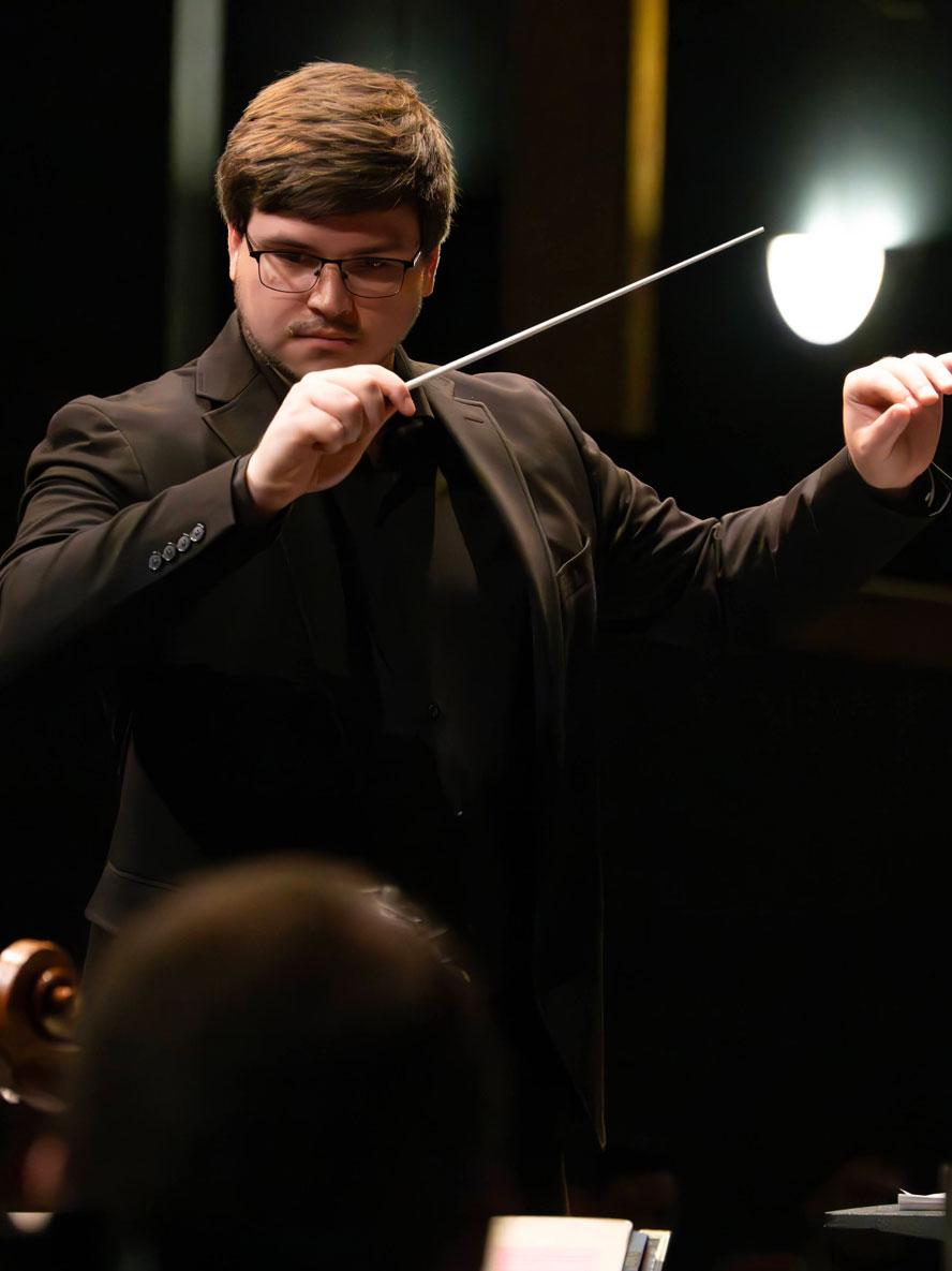 A man with glasses stands and waves a conductor's baton.