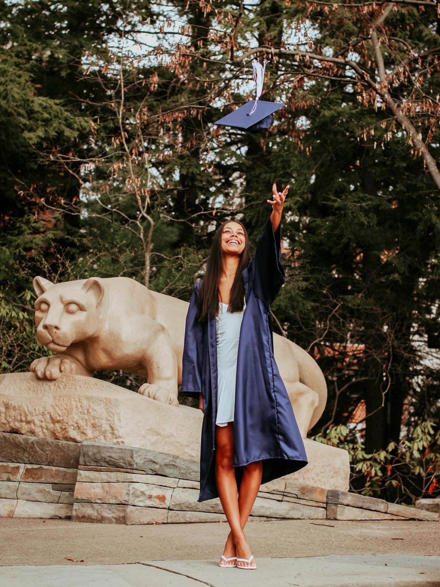 Jazmyn in a blue gown throws her cap in the air near a stone lion statue, surrounded by lush greenery.
