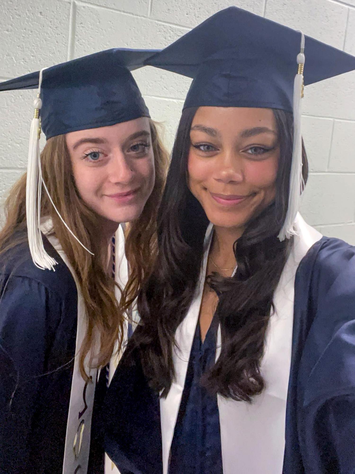 Two graduates wearing navy caps and gowns with white tassels and stoles smile at the camera. The background is a light-colored wall.
