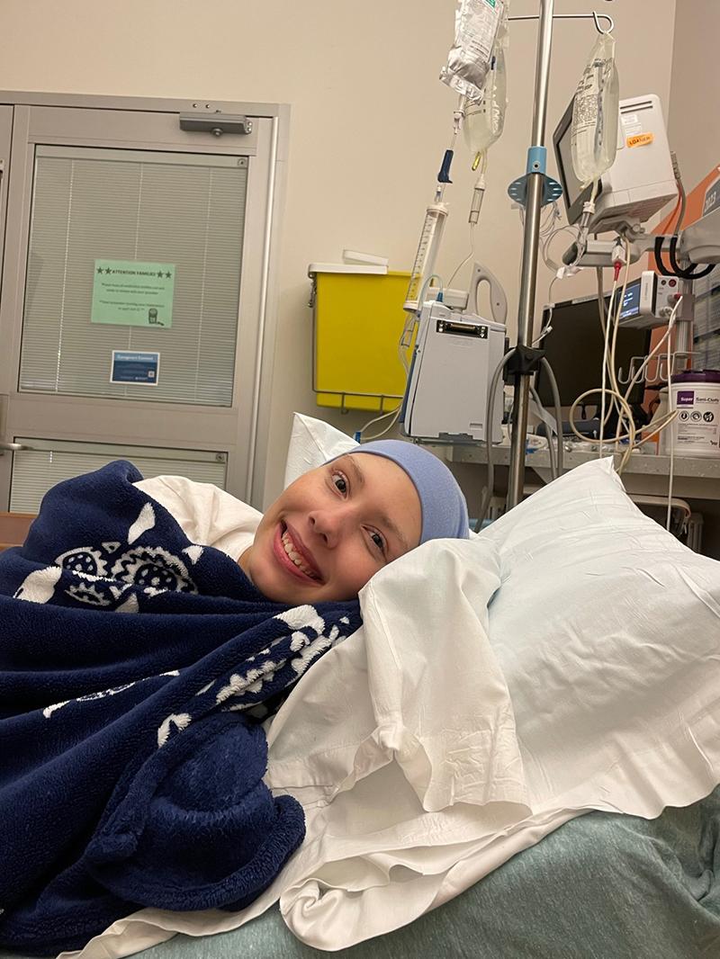 Keira Woods smiles as she lies in bed in a hospital room. She is covered with a blanket and has a tight cap on her head. Behind her on the right is an IV pole with two IV bags and other medical equipment. A door is behind her on the left.