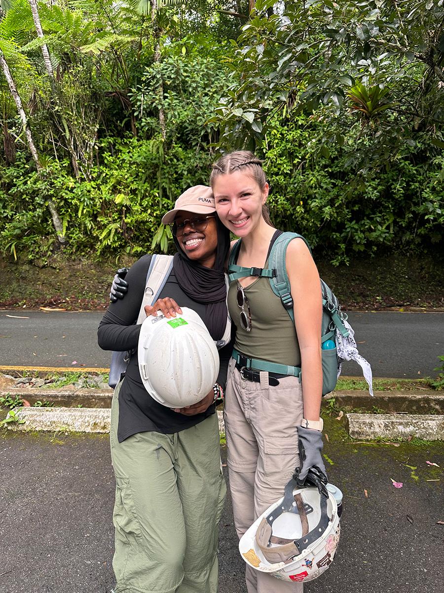 Shannette and Layna Henry stand together outdoors holding hard hats, wearing backpacks and gloves in a lush green setting