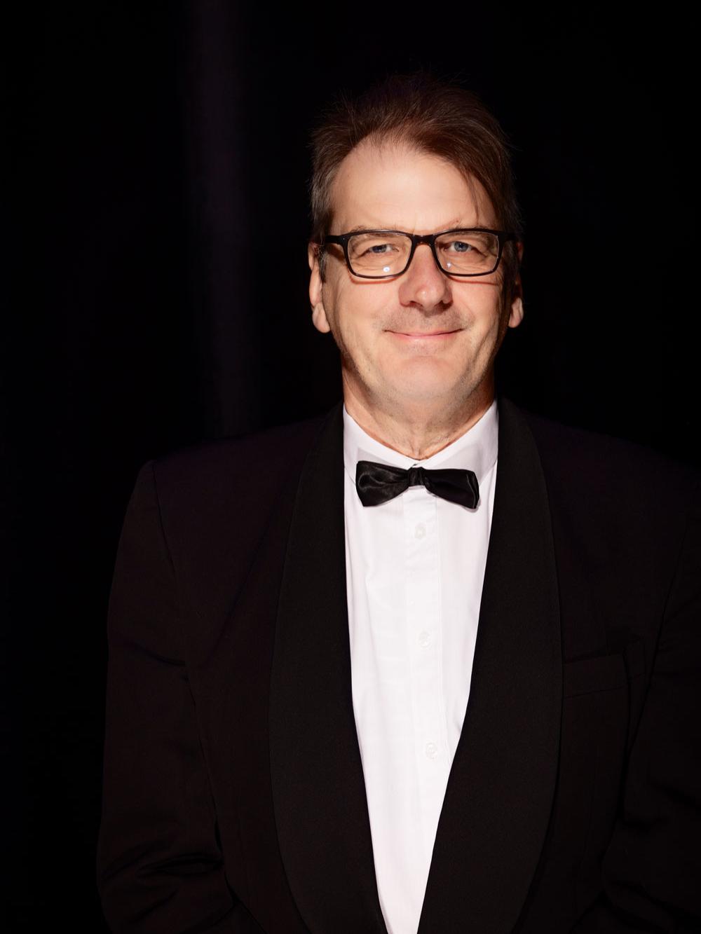 A man wears eyeglasses, bow tie, and tuxedo.