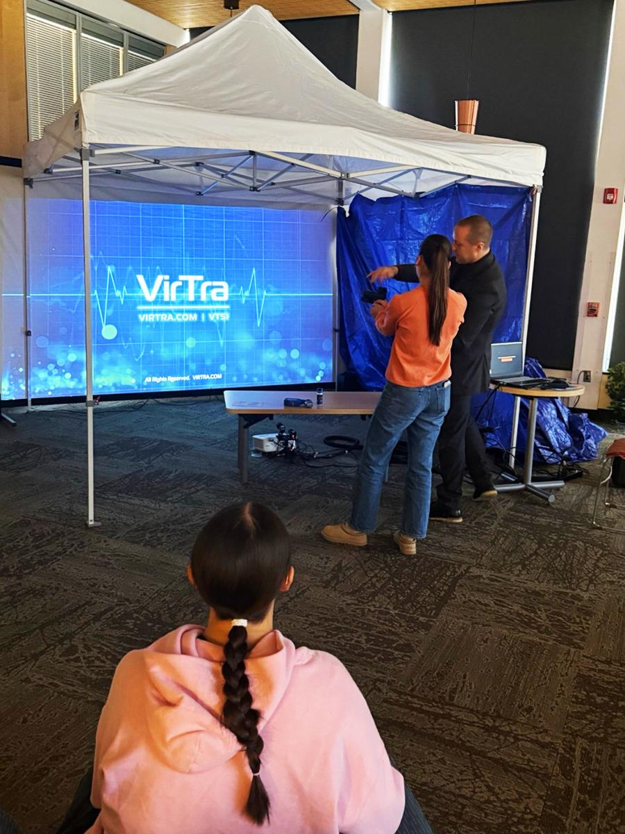 A participant stands under a tented setup practicing a simulated scenario with an instructor, while others observe, with a VirTra training screen displayed in the background.