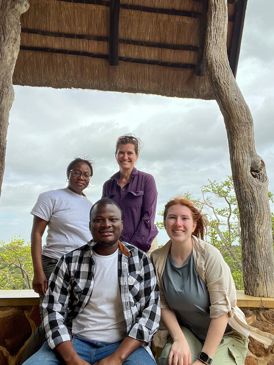 A group of four people smiling under a rustic wooden shelter in a natural setting. The mood is relaxed and joyful, with cloudy skies in the background.