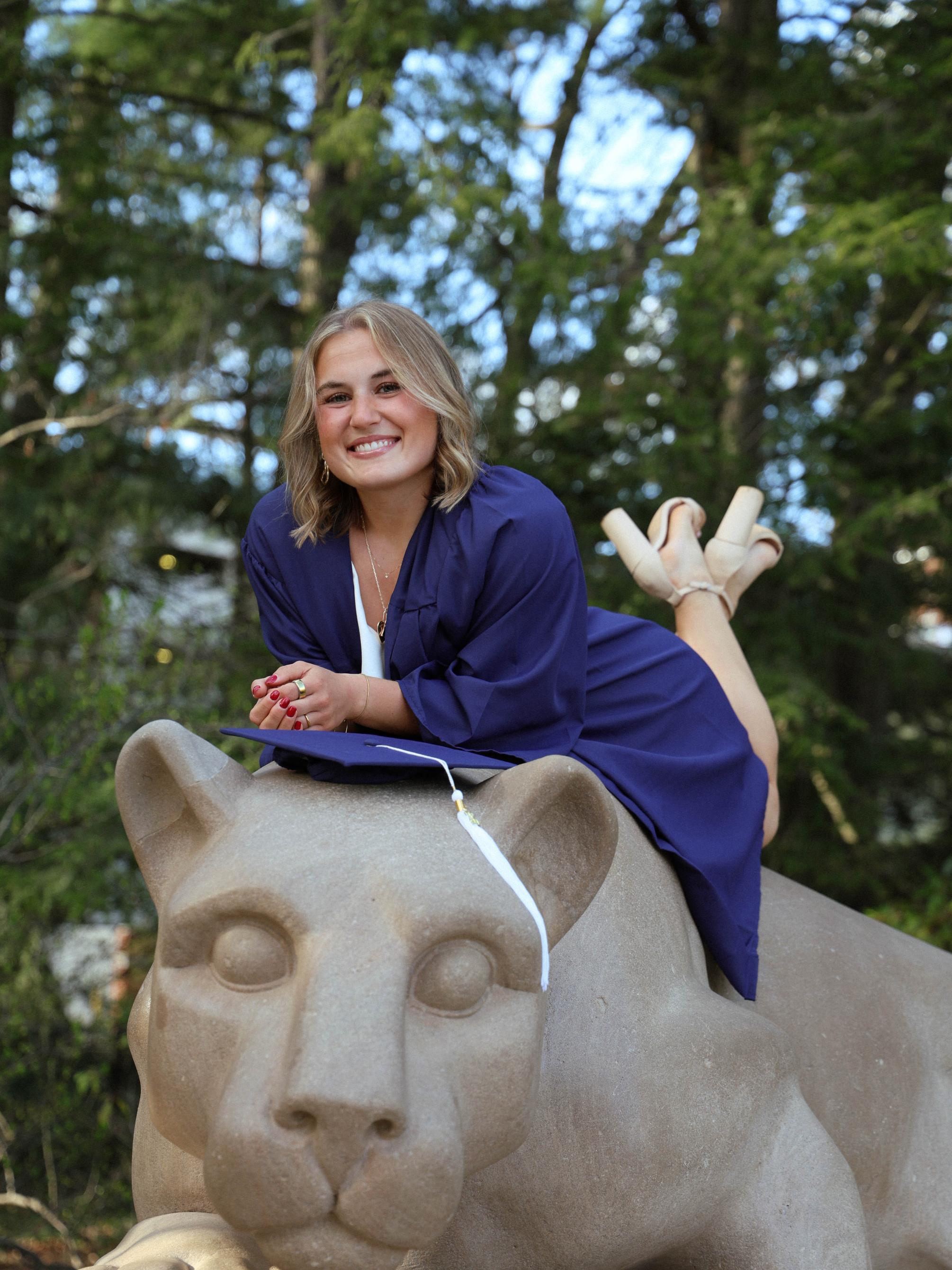 Student in blue graduation gown poses on Nittany Lion sculpture.