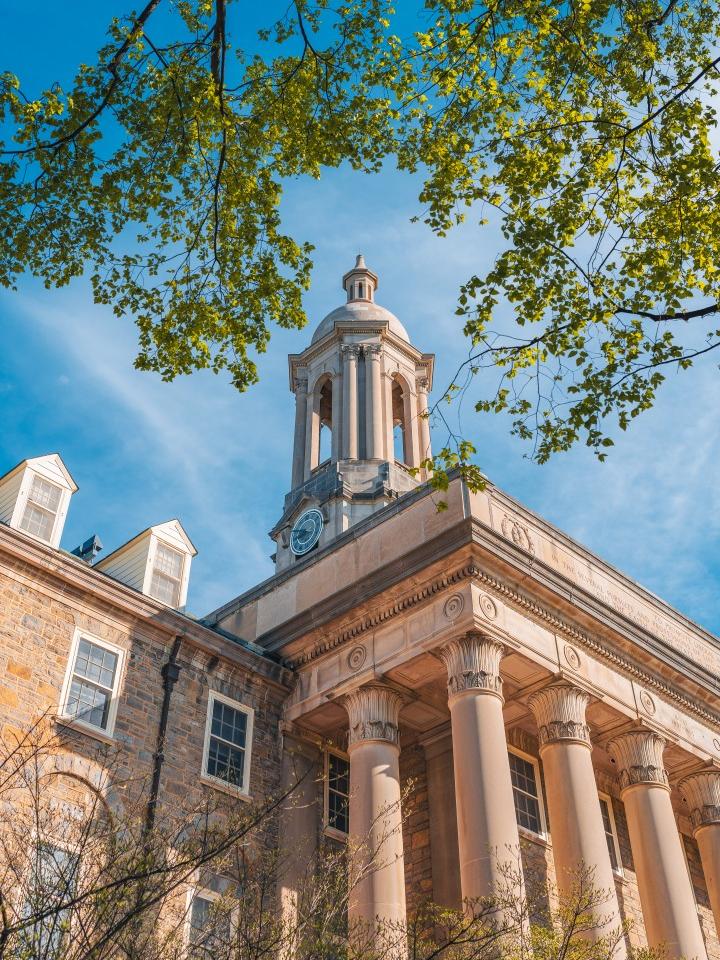 A ground-up shot of the Old Main bell tower with green leaves in the foreground and a blue sky in the background