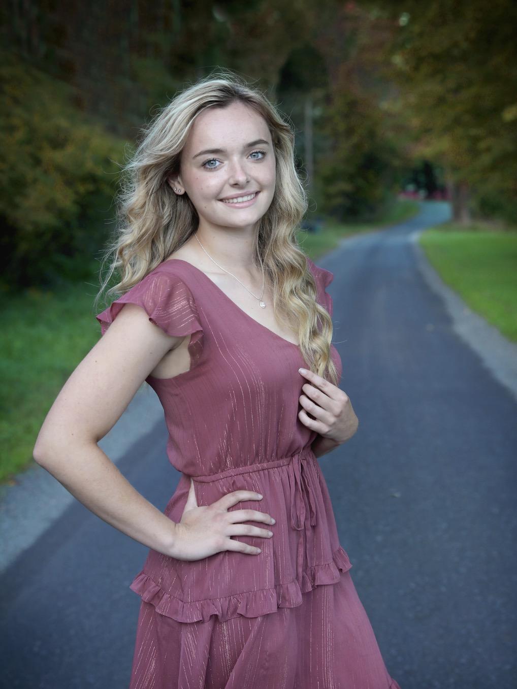 Student in a pink dress posing outside, standing on a paved road in a wooded area.