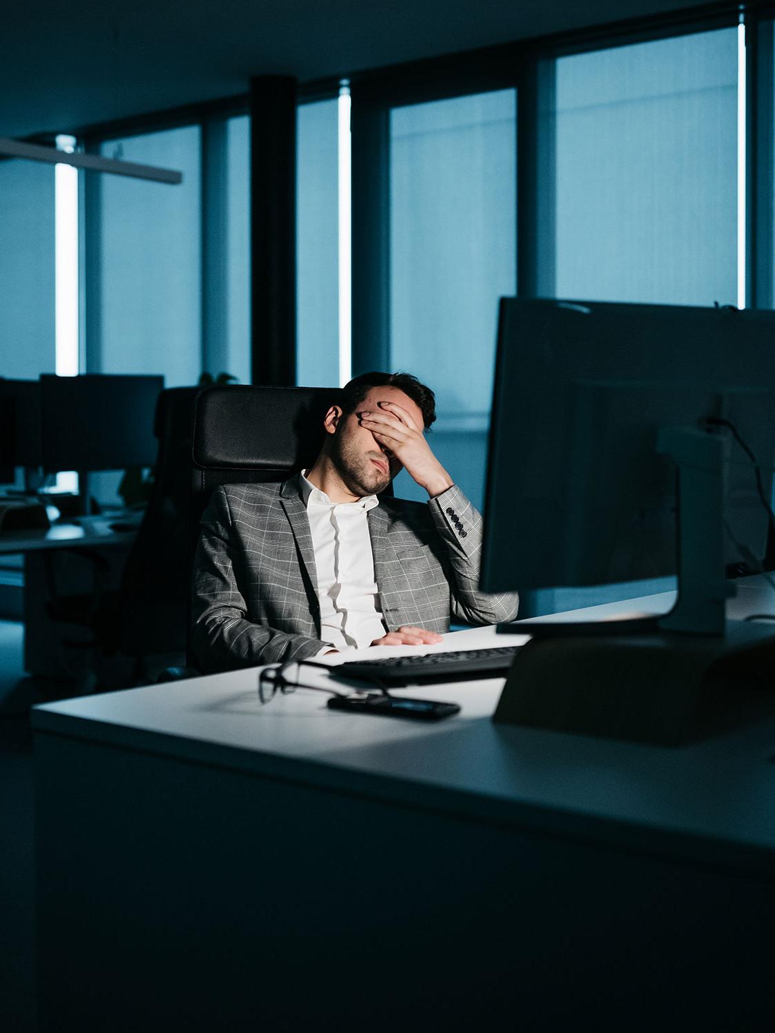 The image shows a smartly dressed businessman resting in his office in front of his computer. He is tired and wants to go home. His face is covered by his hand.