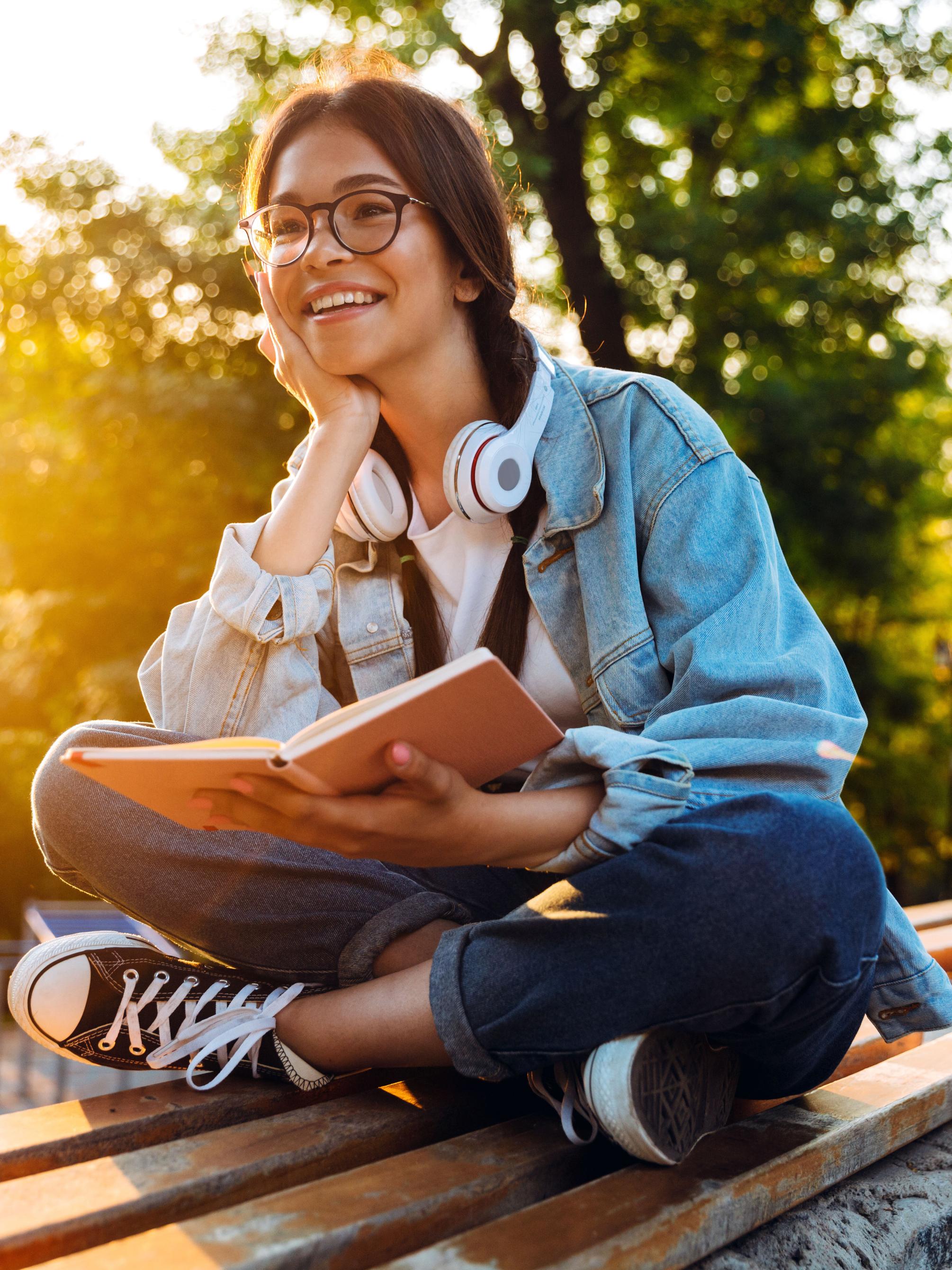 A smiling teenager sits outdoors on a bench reading a book on a sunny summer afternoon.