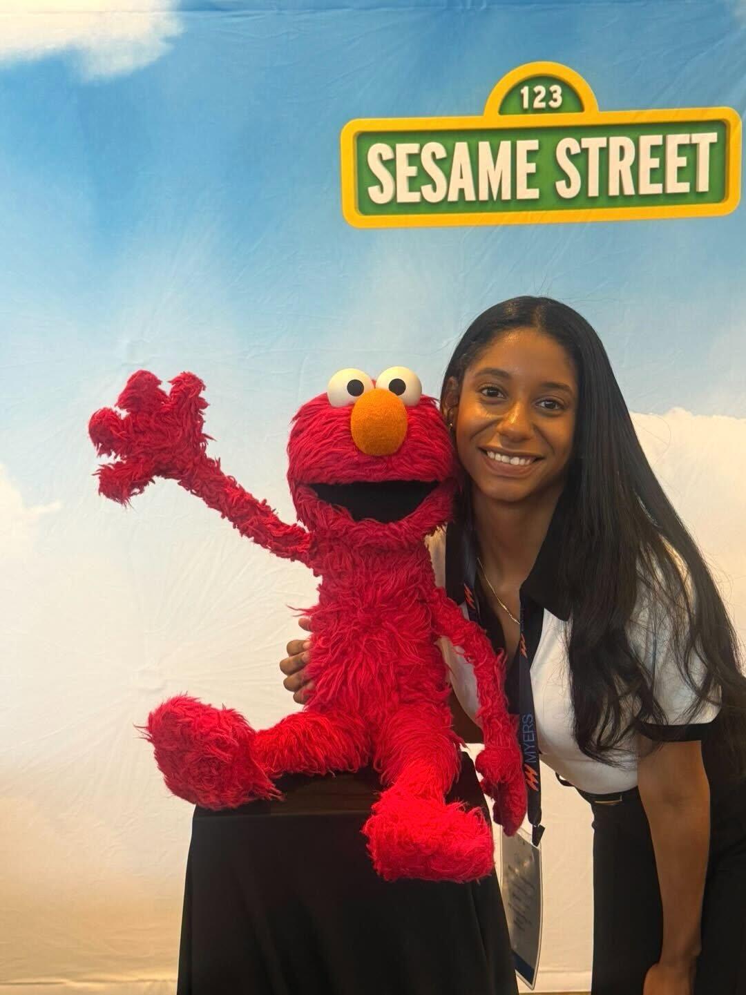 Penn State alumna Claribel Rivas poses in professional attire next to Elmo, a cartoony red puppet, in front of a Sesame Street backdrop