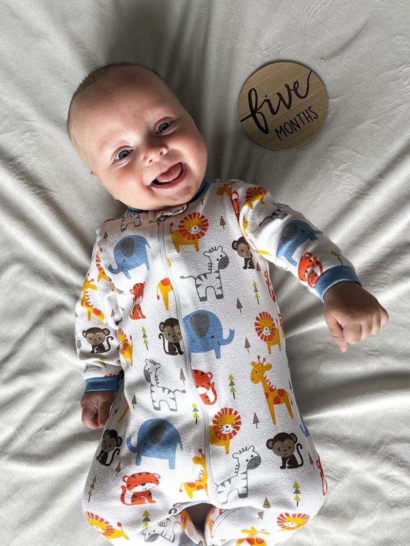 A baby boy smiles as he lies on a blanket wearing a onesie with an animal print. A sign next to him reads “Five months.”