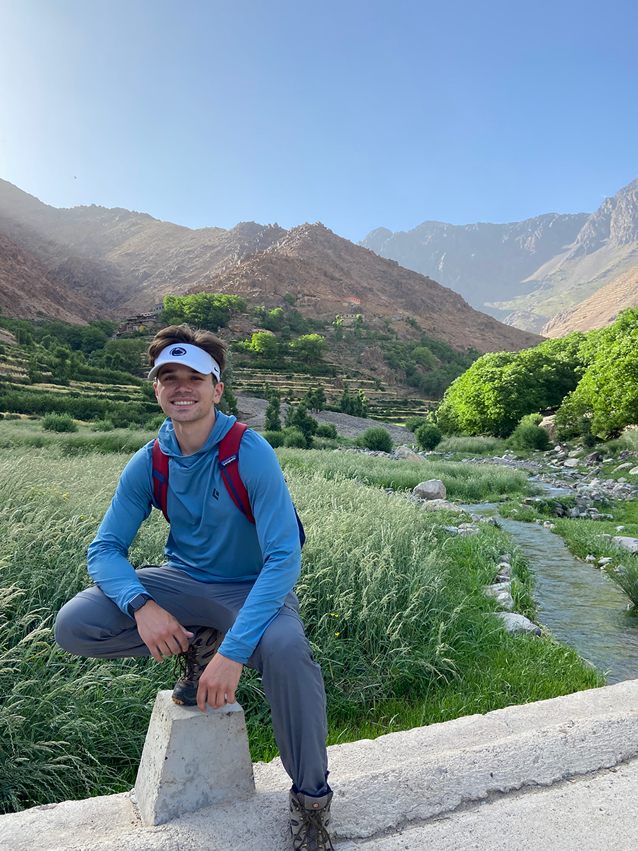 Nate Cherok in a blue long-sleeve shirt and gray pants poses on a concrete block in a mountainous landscape in Morocco, with greenery and a stream in the background.