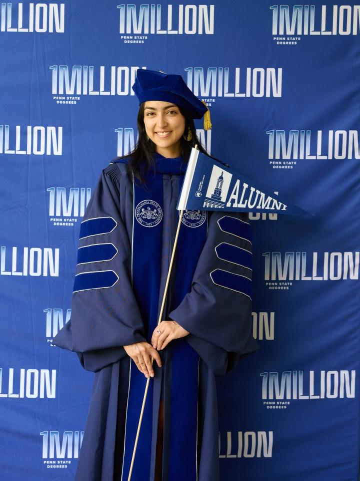 A student wearing regalia poses in front of a backdrop that reads "1MILLION" while holding a pennant that reads "Alumni"