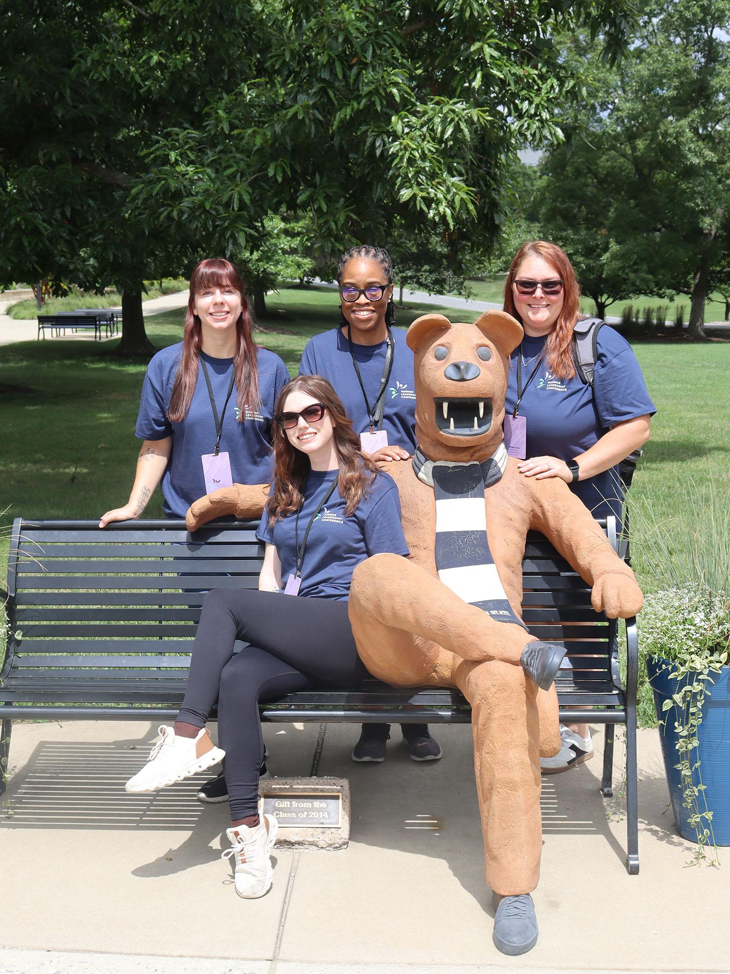 Four women pose with a statue of the Nittany Lion mascot on a bench.