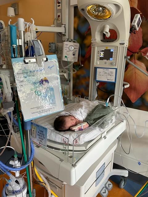 Newborn infant resting in a hospital bassinet within a neonatal care area, surrounded by medical equipment and monitoring devices.