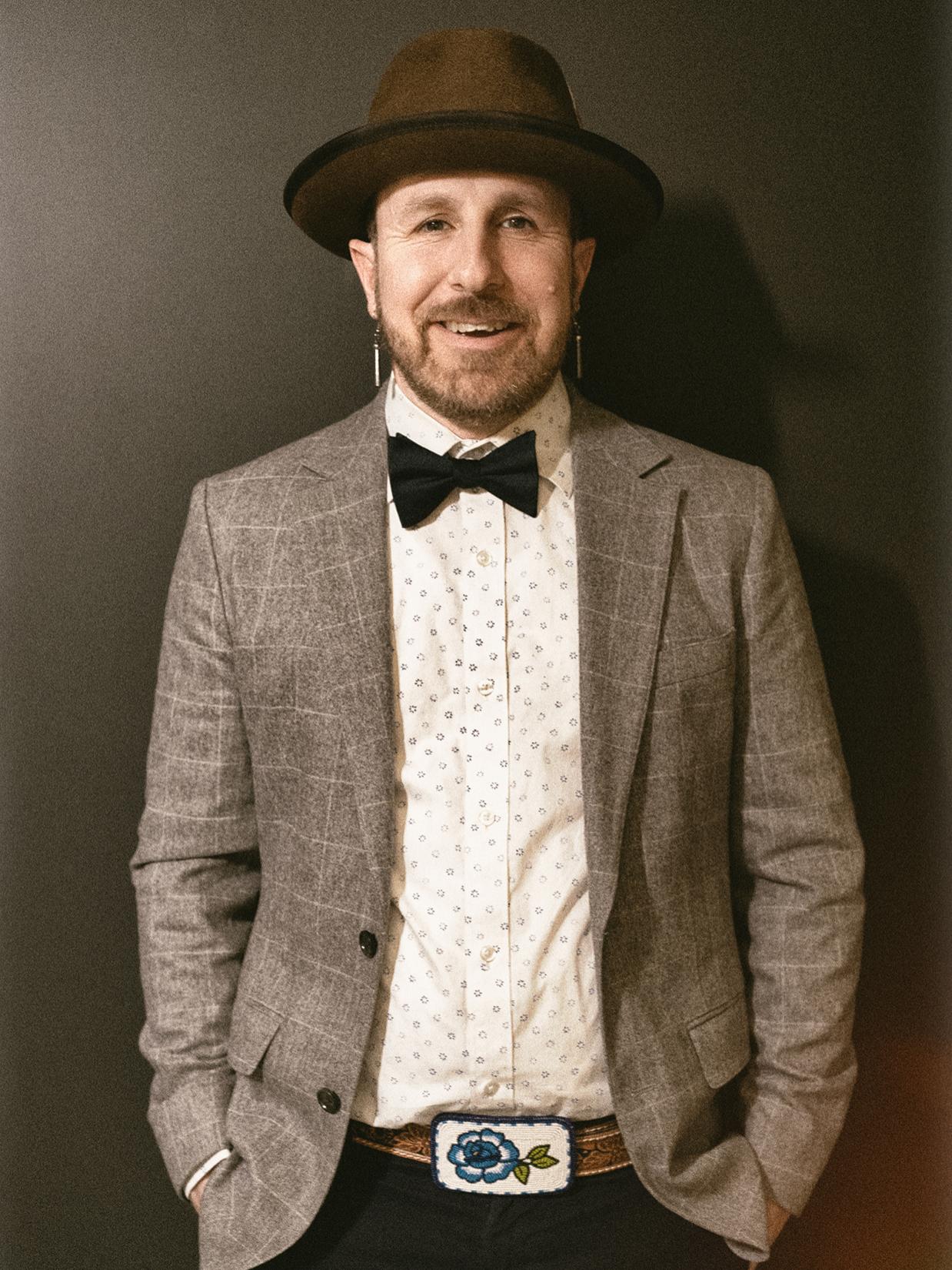 Artist Ryan RedCorn in a brimmed hat, dark bowtie, plaid jacket, and beaded belt buckle, smiling at the camera. He wears long earrings. 