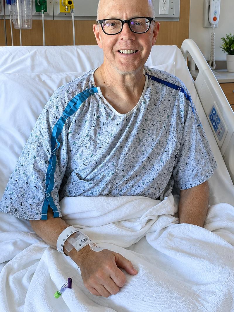 Stephen McNamara smiles from a hospital bed while wearing a hospital gown and holding an IV line.