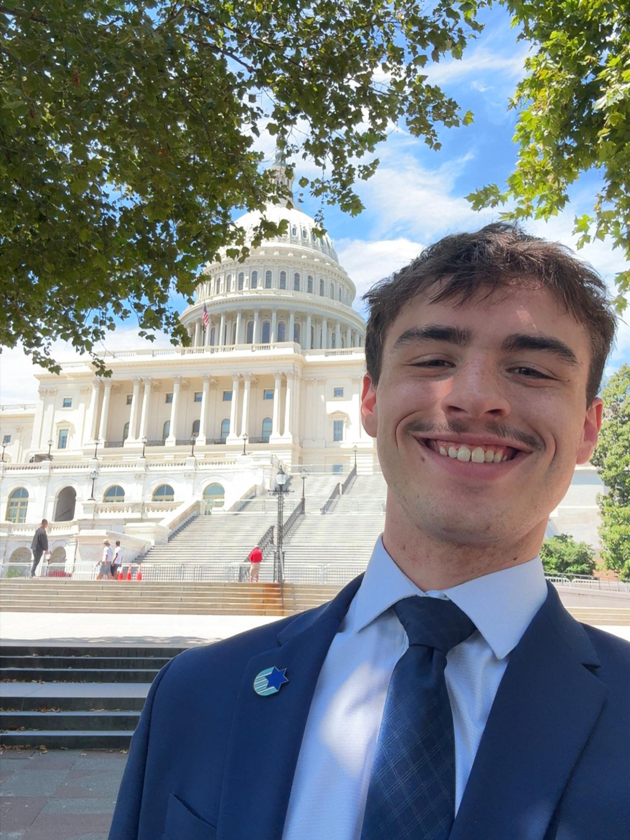 : Dawson Glogau wears a navy suit and tie standing in front of the U.S. Capitol Building. 