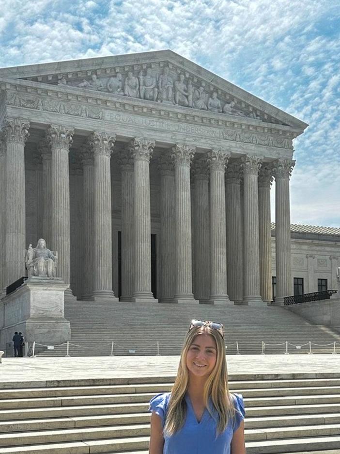 Erin Gregorian in a blue blouse standing in front of the U.S. Supreme Court building on a sunny day with a partly cloudy sky.