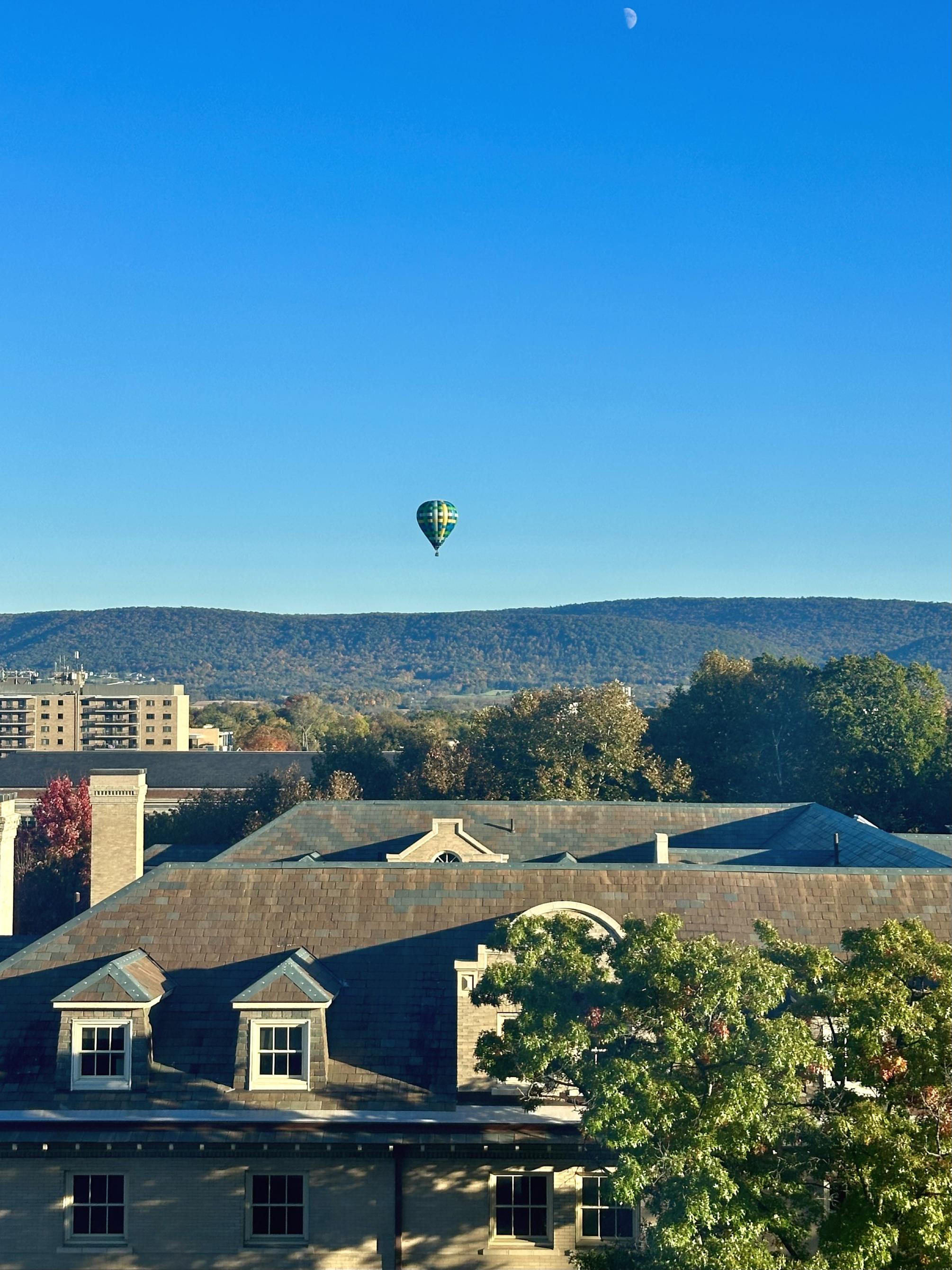 A hot air balloon rises over the blue horizon, with a view of fall forestry and buildings below