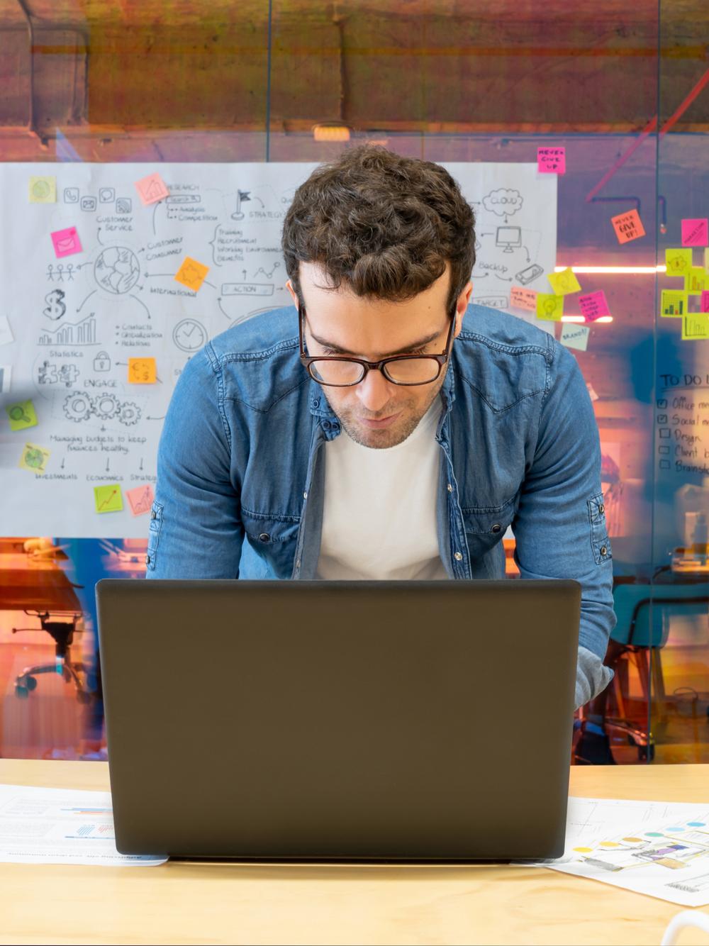Young man with glasses examines a laptop in a bustling office environment