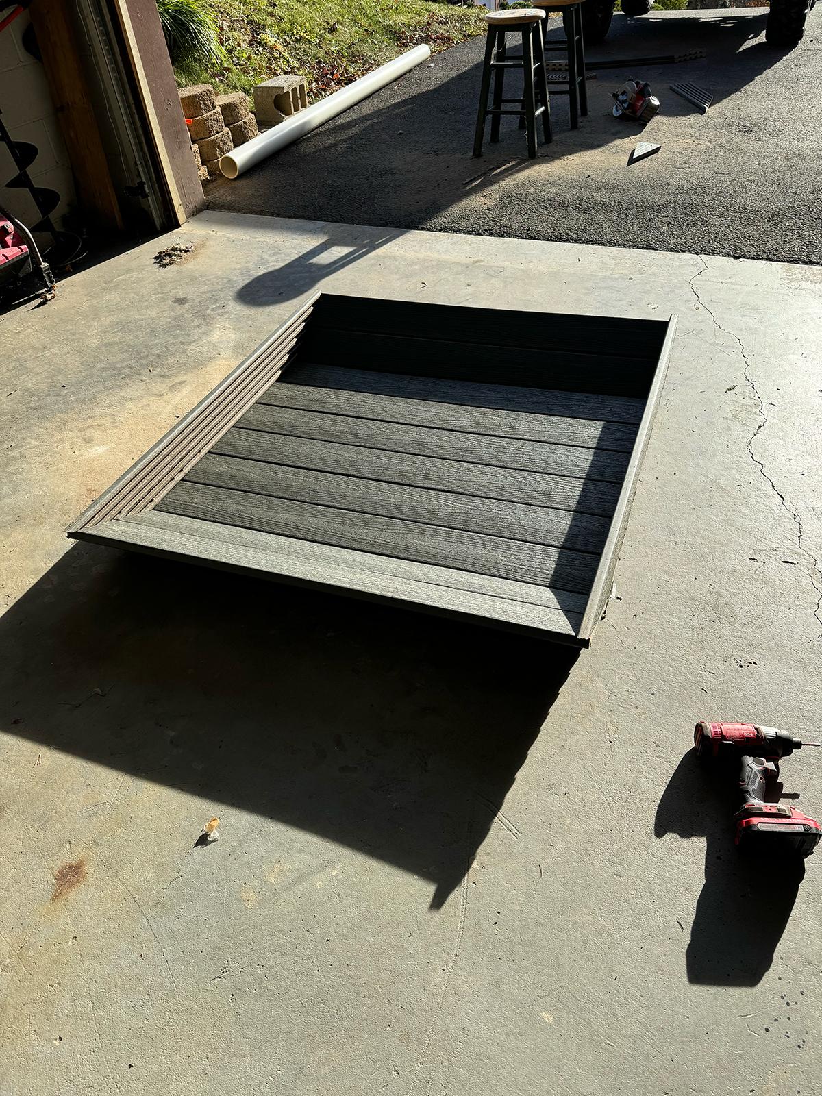 A rectangular wooden platform under construction rests on a concrete driveway, with a cordless drill and construction materials nearby.