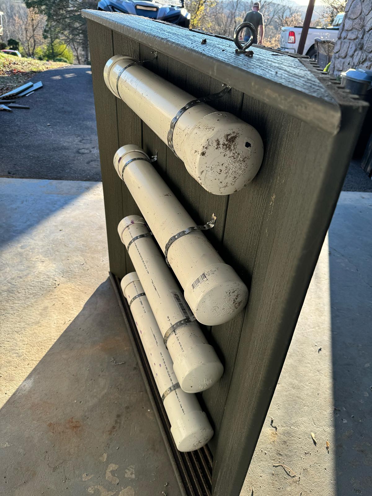 Four white PVC pipes of varying lengths are secured horizontally with metal straps to a dark wooden panel outdoors.