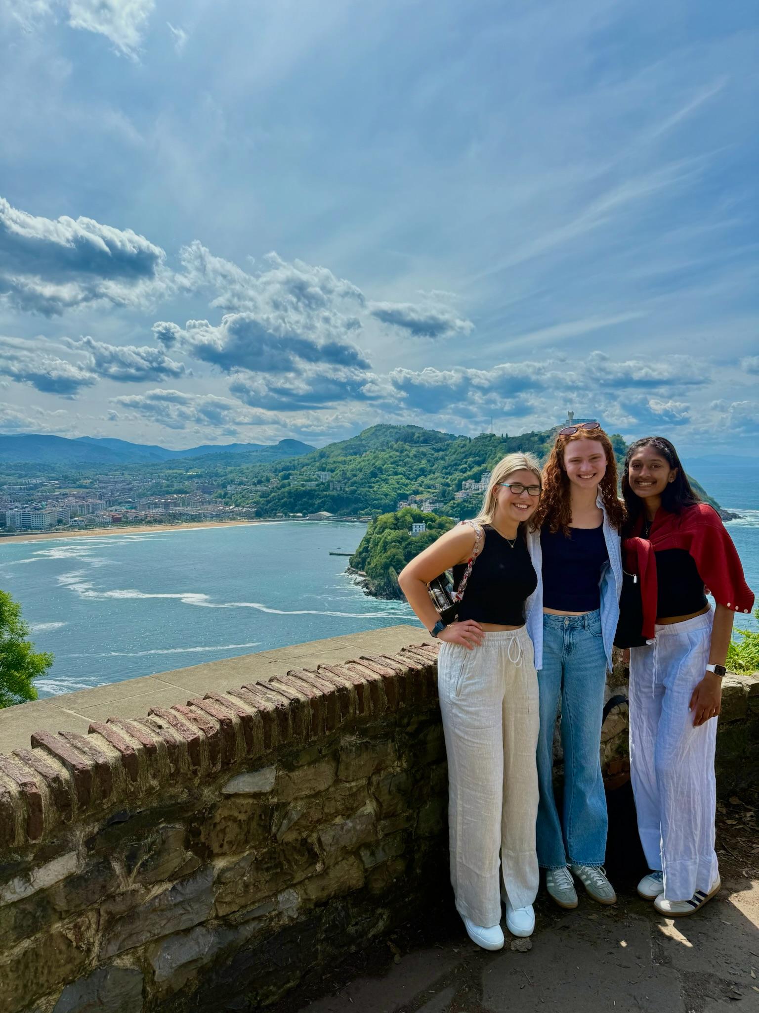 Three young women, smiling and standing in front of a half-wall of stone. The ocean is behind them.