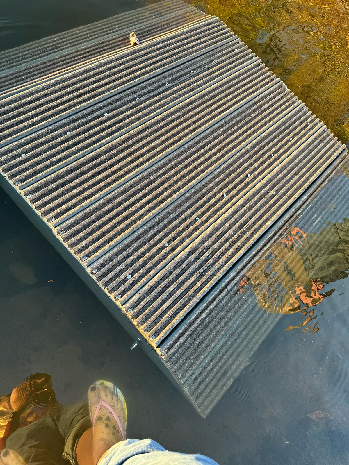 A rectangular, ribbed wooden platform sits partially submerged in water, with autumn leaves floating nearby and a person's feet visible at the bottom.